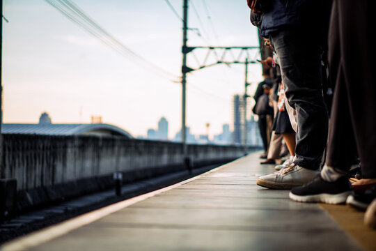 Low angle view of passengers feet waiting on a train station platform at sunset. Blurred city skyline and railway infrastructure in the background. Concept of urban commute and public transport