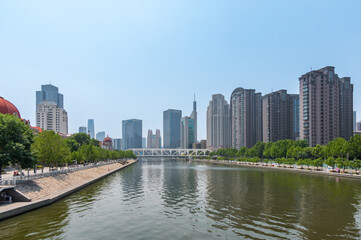 Naklejka premium The Hai River flows through the heart of Tianjin, flanked by a mix of modern skyscrapers and green riverside promenades reflecting in the calm water. China, 22 May 2025