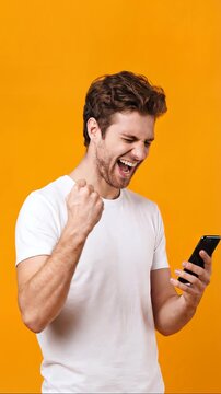 Excited young Caucasian man in a white t-shirt clenches his fist and yells in joy, looking at his smartphone against an orange background. Celebrating good news or online victory.