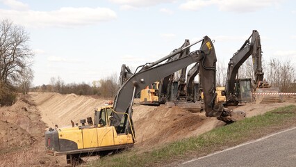 Rząd nowoczesnych koparek gąsienicowych stoi na placu budowy. Potężne, żółte maszyny z długimi ramionami są gotowe do robót ziemnych. W tle widać drzewa i błękitne niebo z delikatnymi chmurami © tomo81