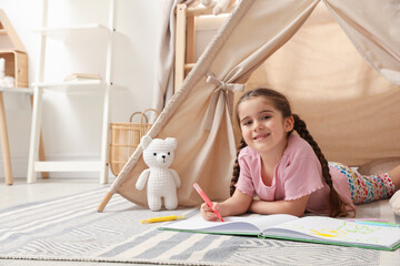 Little girl drawing in sketchbook near toy wigwam at home, space for text © New Africa