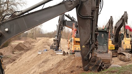 Rząd nowoczesnych koparek gąsienicowych stoi na placu budowy. Potężne, żółte maszyny z długimi ramionami są gotowe do robót ziemnych. W tle widać drzewa i błękitne niebo z delikatnymi chmurami © tomo81