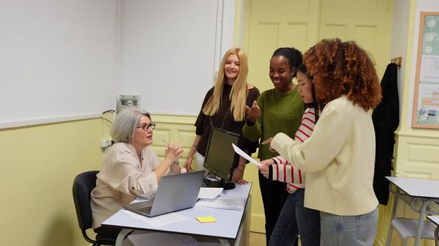 Mature female teacher explaining schoolwork to a multiethnic group of young university students gathered around her desk in a classroom
