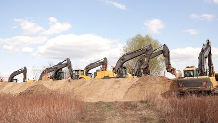 Rząd nowoczesnych koparek gąsienicowych stoi na placu budowy. Potężne, żółte maszyny z długimi ramionami są gotowe do robót ziemnych. W tle widać drzewa i błękitne niebo z delikatnymi chmurami © tomo81