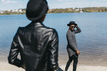 Fototapeta premium African American couple enjoying outdoors by lake, man looking back at woman, wearing stylish hats and urban fashion