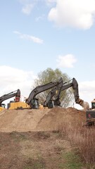 Rząd nowoczesnych koparek gąsienicowych stoi na placu budowy. Potężne, żółte maszyny z długimi ramionami są gotowe do robót ziemnych. W tle widać drzewa i błękitne niebo z delikatnymi chmurami © tomo81