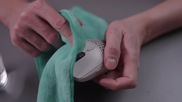 Person cleaning white wireless gaming computer mouse with microfiber cloth, disinfecting electronic peripheral device for hygiene and maintenance on desk, close-up. Shooting in slow motion.