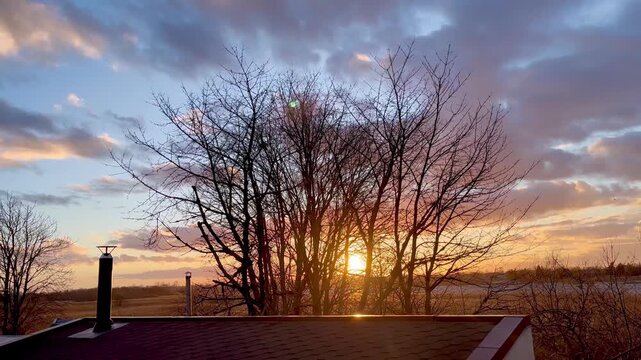 Sunset shining through leafless trees above house roof and chimney with colorful evening sky over rural landscape during calm early spring countryside scene