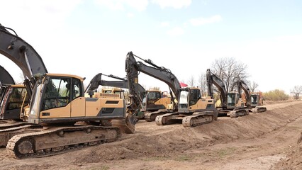 Rząd nowoczesnych koparek gąsienicowych stoi na placu budowy. Potężne, żółte maszyny z długimi ramionami są gotowe do robót ziemnych. W tle widać drzewa i błękitne niebo z delikatnymi chmurami © tomo81