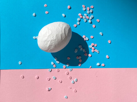 White egg with colorful confetti on blue and pink background. High angle view, minimalist composition.A textured white chicken egg lies on a vibrant dual-tone blue and pink paper background.
