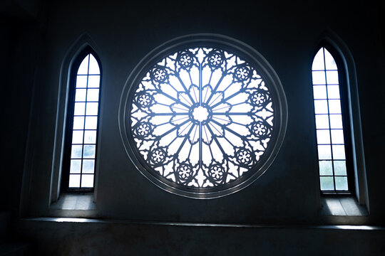 Beautiful Ornate circular stained glass style window inside a dark Gothic church interior between two pointed cathedral windows.