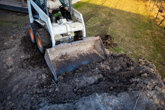 A worker operates a white skid steer with bucket, pushing wet soil on a sloped lawn beside a house deck. Mud caked tires, operator cage, and clear cut earth are visible.