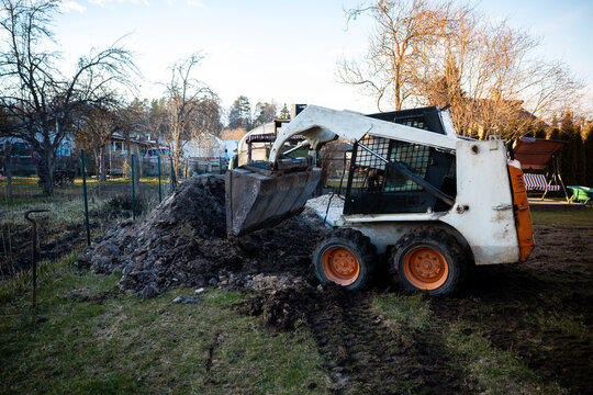 Compact skid steer loader lifts wet soil beside a fenced yard, mud splattered bucket mid lift in a tight residential setting, cool late day light and long shadows