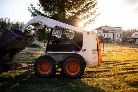 A compact skid steer with orange wheels dumps dark soil on rutted grass as warm late sun flares through a conifer near cinder block walls and scaffolding.