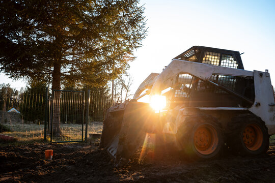 A compact skid steer with front bucket sits on churned soil at dawn. Backlighting casts flares, mud marks the white body and orange wheels, work prep is underway.