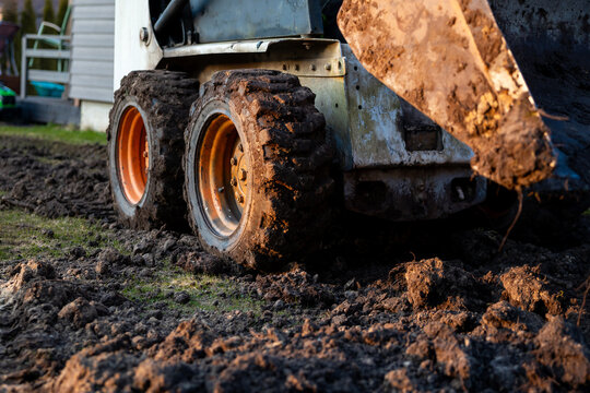 Ground level view of a compact skid steer with bucket grading wet rutted soil, mud on thick tread tires, siding wall and garden ladder suggest yard drainage work.