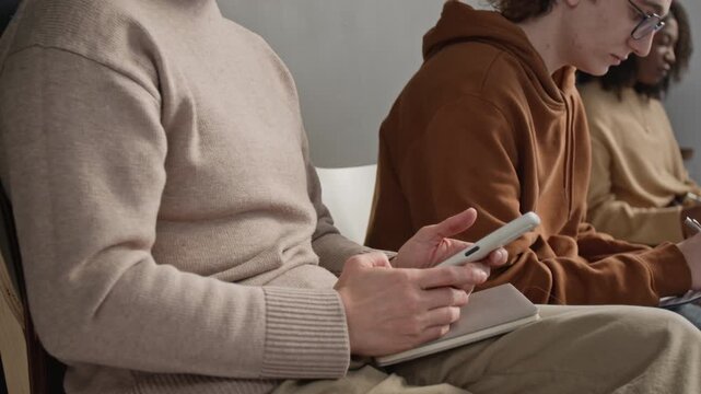 Medium shot of mature adult Caucasian woman scrolling social media on smartphone while sitting with other patients in hospital waiting room
