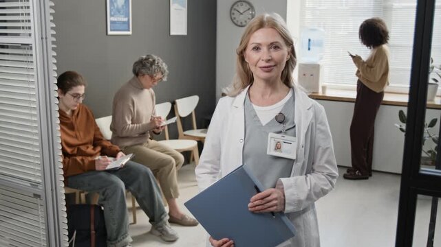 Medium portrait of confident senior Caucasian female medical worker holding file folder smiling at camera, diverse patients visible at waiting area in background