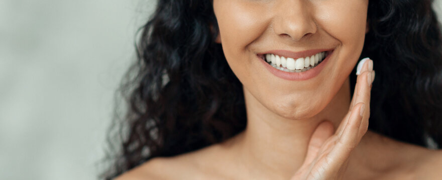 A woman with curly hair smiles while placing her hand on her cheek. She appears happy and relaxed. Soft light highlights her features, showing a clear complexion and bright smile.