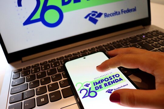 Salvador, Brazil - April 13, 2026: Woman's hand holding a smartphone with Declara&ccedil;&atilde;o do Imposto de Renda 2026 logo (Brazil Income Tax Declaration 2026) and a laptop in the background. 