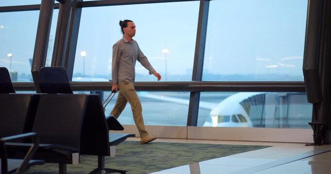 Slow motion full-length shot of passenger man in casual attire walking with trolley suitcase through modern airport terminal. Airplane is visible through large glass window in background