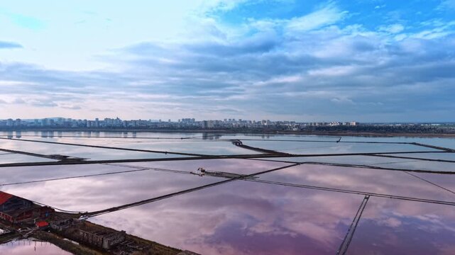 Moving over the dilapidated houses at the waterfront. Drone flight above the lakes used for salt production. City skyline under cloudy sky in the backdrop.
