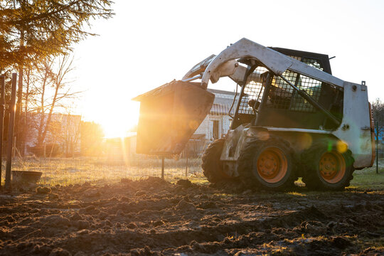 A compact skid steer raises a front bucket beside a wire fence as low sun backlights metal mesh and orange wheels, with lens flare and long shadows on mud rutted ground.