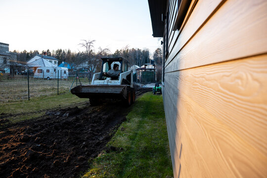 Compact track loader levels muddy soil beside modern wood clad house, leaving deep ruts next to sunlit siding in late afternoon, with fenced lots and outbuildings nearby.