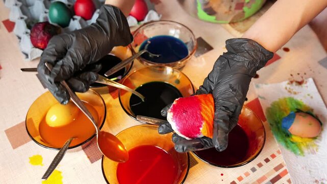 Close-up video of gloved hand holding an egg wrapped in a napkin while colored dyes are poured with a spoon, creating a multicolored Easter pattern. 2026