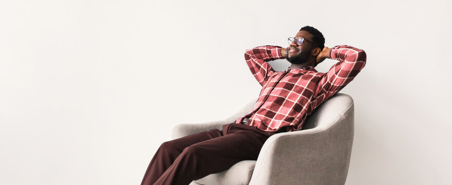 A man sits in a modern chair with a relaxed posture. He has his hands behind his head and smiles while enjoying a moment of leisure. The setting features simple and light colors.
