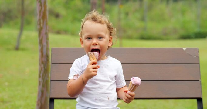 Cheerful young boy sits on park bench, indulging in joy of double treat - holding two ice cream cones. His happiness is evident as he licks and enjoys tasty frozen treats.