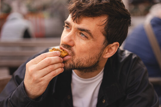 Close up of hungry middle-aged man with blue eyes and thoughtful facial expression eating delicious cookie or pastry outdoors, handsome male in black jacket enjoying sweet snack at street cafe