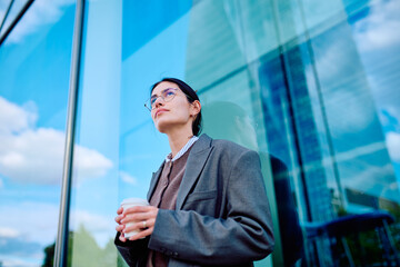 Young businesswoman looking up, holding a coffee cup, standing next to a modern glass building with reflections of the sky © we.bond.creations