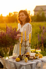 Fototapeta premium Gorgeous beautiful woman in a white sundress enjoying picnic in a picturesque place. Romantic date, marriage proposal, engagement. Fancy elegant table setting in a violet lupins field or meadow