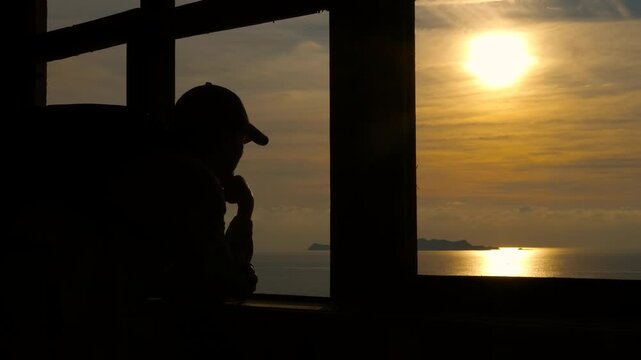 Pensive silhouette of a man watching the sunset over the ocean. Silhouette of a pensive man wearing a baseball cap, looking out a window at a beautiful golden sunset over the tranquil ocean