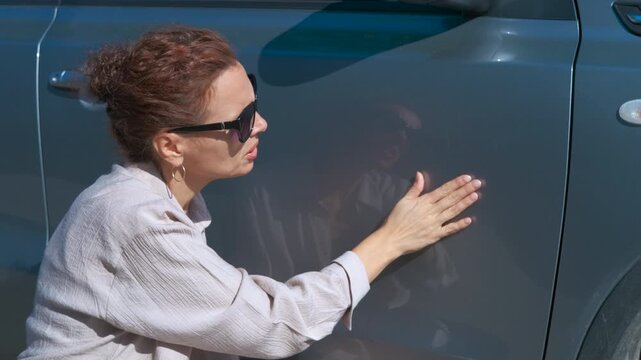 Woman inspecting car for scratches and dents. Meticulous redhead driver wearing sunglasses carefully examining gray car exterior, checking for potential scratches, dents, and surface imperfections
