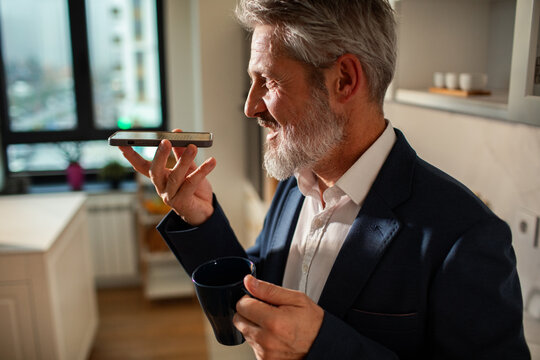 Middle-aged businessman using voice assistant on smartphone at home kitchen