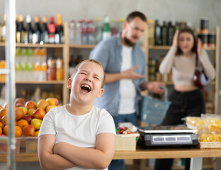 Fototapeta premium Capricious preteen boy standing with crossed arms and screaming loudly in grocery store while frustrated puzzled parents looking at son in background ..