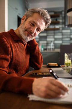 Smiling mature man writing notes at home kitchen table