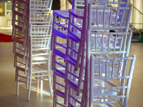Rows of elegant white wooden chiavari chairs stacked vertically in banquet hall under vibrant purple and warm ambient lighting following professional celebration or wedding reception.