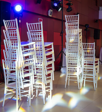 Vertical stacks of elegant white wooden chiavari chairs standing in empty event space illuminated by professional blue and yellow stage spotlights against red paneled wall.