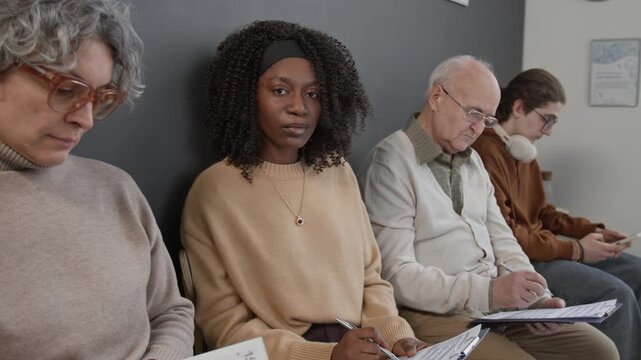 Medium shot of young Black woman holding clipboard with medical form looking at camera while waiting for doctor in queue
