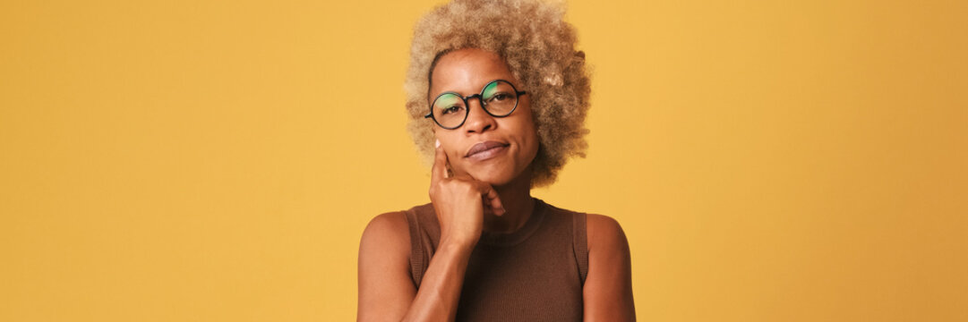 Beautiful girl in glasses, wearing brown top listens attentively to someone and disagrees isolated on orange background. Panoramic