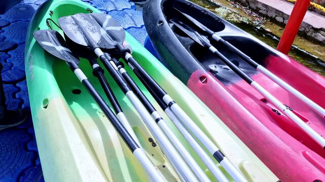 A colorful dockside scene in Nainital, Uttarakhand, India, showing neatly stacked paddles resting across green and red kayaks placed on a textured floating platform by lakeside.