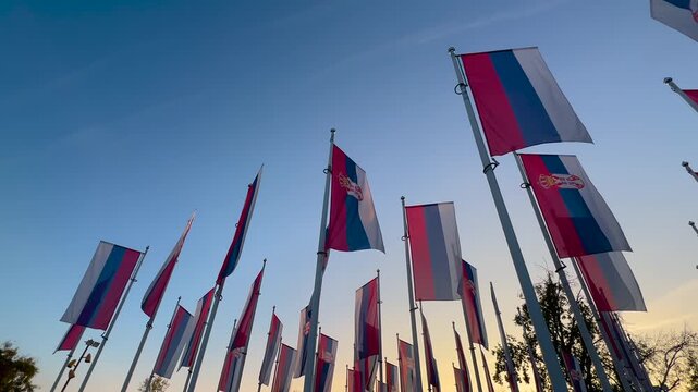 Multiple Serbian national flags on tall flagpoles waving in the wind against evening sky during sunset in Novi Sad, Serbia, representing national identity, patriotism and public space