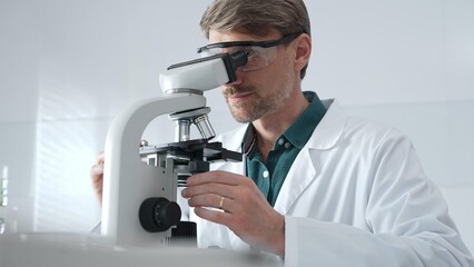 Adult male scientist wearing lab coat and protective glasses using microscope, doing scientific...