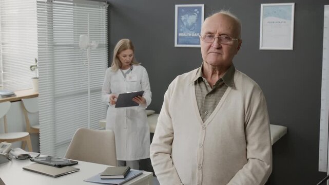 Medium portrait of senior Caucasian man wearing eyeglasses standing at doctors office in office, female physician holding clipboard looking through records in background