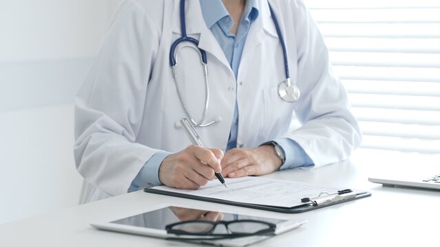 Female doctor documents patient information while working with digital devices at minimalist white workstation in medical office. Medicine and healthcare concept