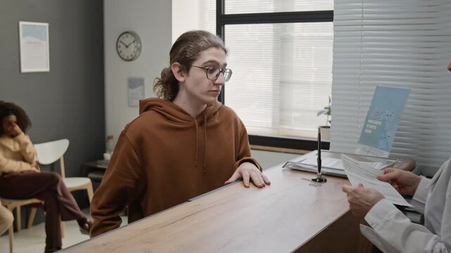 Teenage boy standing at reception desk showing appointment information in smartphone app, mature female receptionist handing him medical paper form