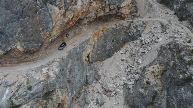 Aerial view of car driving along extremely dangerous rocky mountain cliff road track. Rugged vehicle follows a perilous ledge route above steep stone slopes in remote Himalaya.